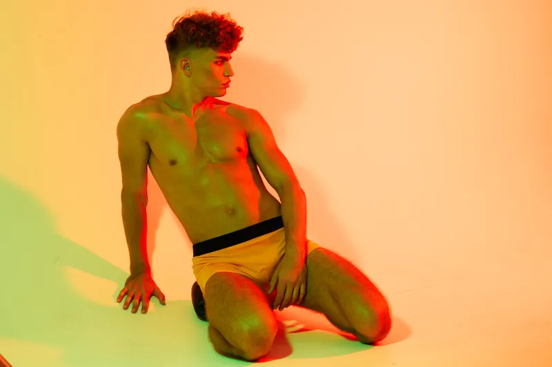 Young man with muscular body posing shirtless on knees against orange background with green and red lighting for fitness, health and fashion concept in studio.