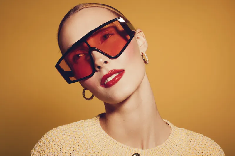 Stylish woman with short hair wearing oversized red sunglasses and gold hoop earrings poses against a warm yellow background. Fashion concept with bold accessories and makeup.