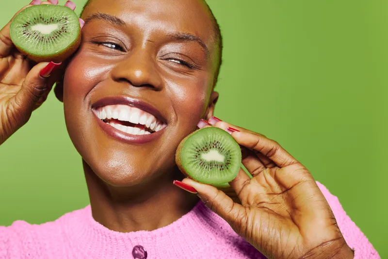 Smiling dark skinned African woman holds two halves of fresh kiwi near face on green background. Concept of healthy lifestyle, fruit, natural beauty, and joyful mood in studio portrait.