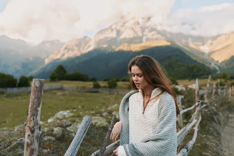 A woman in a cozy sweater stands by a weathered fence in a mountainous landscape, embodying a calm, outdoorsy lifestyle moment.