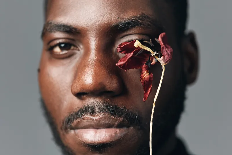 Creative conceptual portrait of African man with dark skin holding dried red flower near eye in studio. Closeup face with artistic visual style and emotional expression for modern photography.