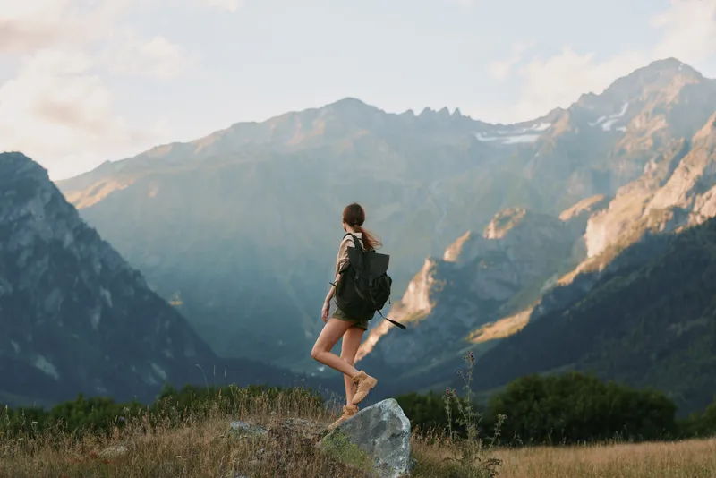 A young woman with a backpack leaps on a rock in a mountain meadow, conveying a vivid sense of outdoor adventure, exploration, and freedom amid towering peaks.