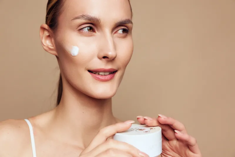 Woman with skincare face cream moisturizer for beauty, close up spa routine, fresh smooth skin, soft natural light, calm expression, hands holding cream jar, minimal beige background