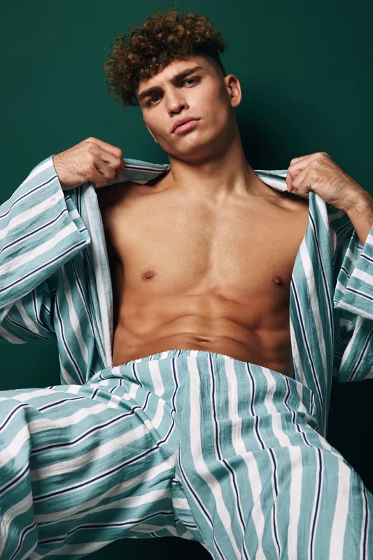 Young man with curly hair wearing striped pajamas showing muscular torso and abs sitting against dark background in studio. Casual fashion and confident pose for lifestyle portrait.
