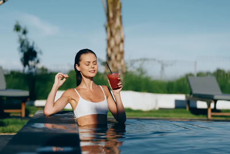 Young woman in white bikini relaxing in outdoor pool holding red cocktail with straw enjoying sunny day with closed eyes and peaceful expression near palm tree and lounge chairs.