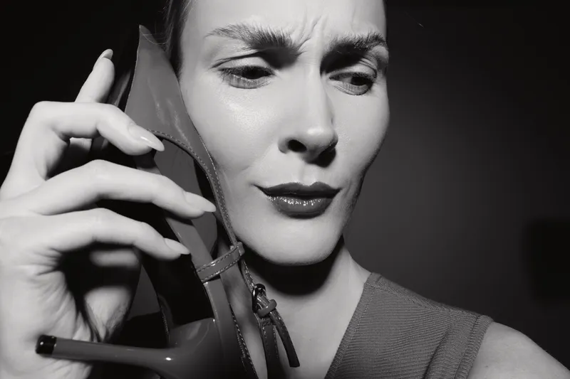 Caucasian woman with fair skin holds high heel shoe near face with confused expression. Black and white studio portrait with dramatic lighting and fashion concept.