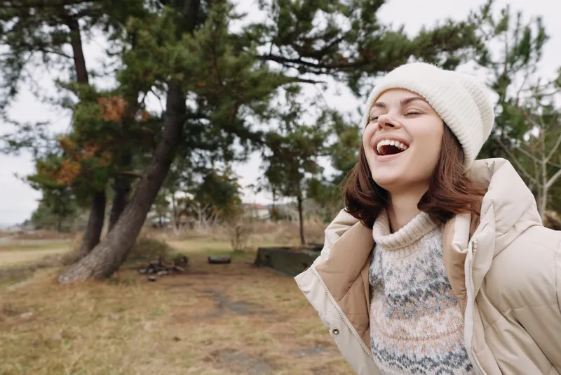 Woman enjoying an outdoor winter moment, laughing with a beanie and puffer jacket in a park, trees and open space around, bright mood and carefree energy.