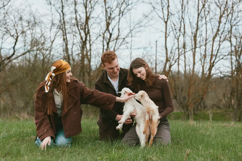 Three friends interact with a playful goat kid in a park, outdoors, sharing a joyful lifestyle moment, animal affection and casual smiles, candid nature scene
