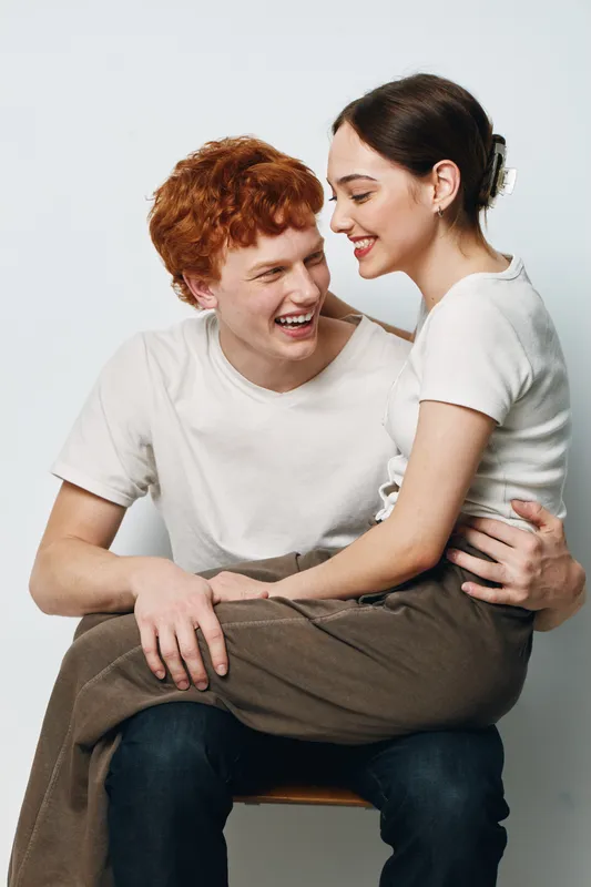 Couple in casual clothing smiling and embracing, young man with red hair sitting on chair and woman in white top sitting on his lap with happy expression in studio.