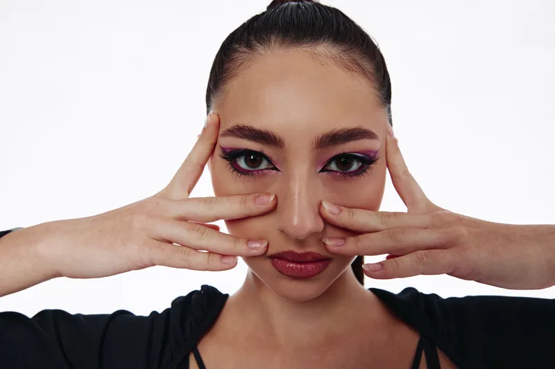 Creative woman with sharply styled hair poses with fingers on face, showcasing dramatic pink eyeliner and glossy lips. Fashion beauty concept on a white background.