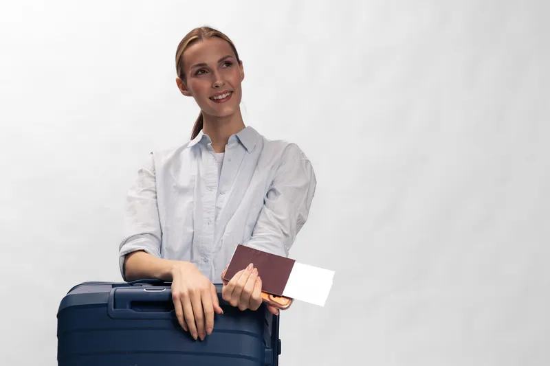 Young smiling woman holding passport and ticket stands with suitcase on white background. Concept for travel, vacation, tourism, and holiday preparation.