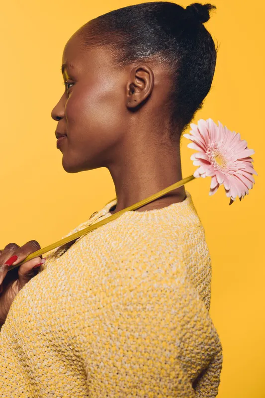 Profile of a smiling black African woman with dark skin holding a pink flower behind her shoulder. Yellow background and knitted sweater create warm, bright, positive mood concept of nature.