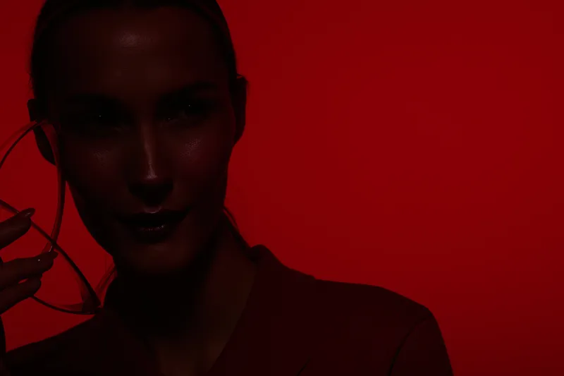 Portrait of a woman with dark skin in red lighting holding eyeglasses near her face. Concept of fashion, beauty, and dramatic mood with strong red background.