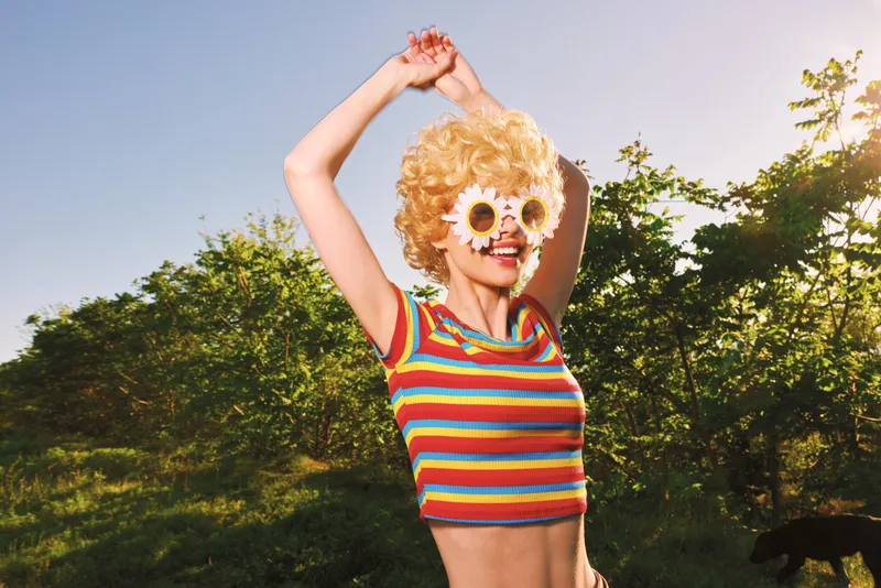 Vibrant woman outdoors wearing a striped top and flower sunglasses, expressing carefree joy in bright sunlight, retro film color mood, and warm summer lifestyle. soft lighting adds warmth and a