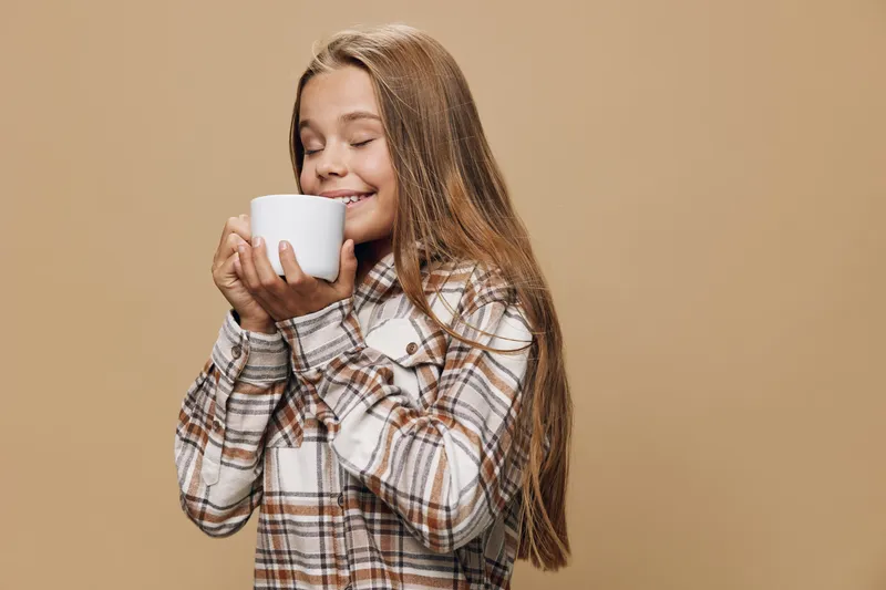 Girl holding white cup with closed eyes enjoying warm beverage in cozy plaid shirt on beige background for comfort and relaxation at home.
