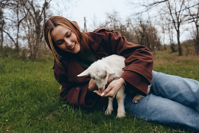Young woman with braided hair lies on grass smiling as she gently feeds a small white baby goat in a natural outdoor setting during a calm autumn day.