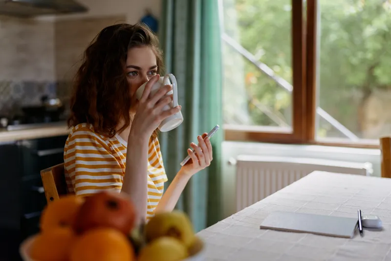 Woman in striped shirt enjoys a quiet morning at home, sipping from a mug, seated beside a fruit bowl near a sunlit kitchen window, conveying calm casual lifestyle