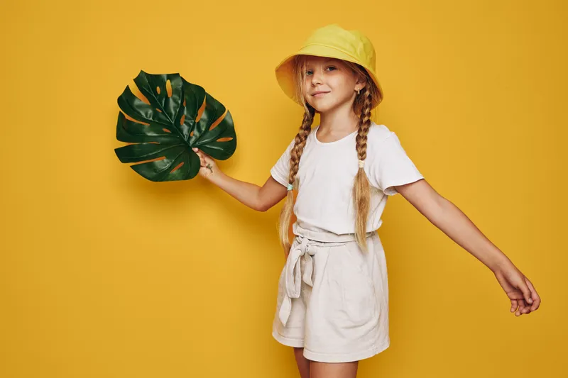 Smiling girl in a yellow hat holds a large tropical leaf against a yellow background. She wears light summer clothes.