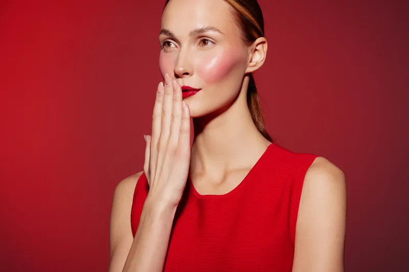 Young woman in red dress with red lipstick covers mouth with hand on red background. Concept of surprise, secrecy, emotion, and fashion portrait with studio lighting.