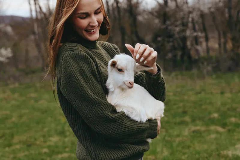 Smiling woman in a knit sweater holds a friendly goat kid in an open field, warm natural light enhancing a tender moment. Lifestyle scene showing companionship, nature, and everyday farming joy.