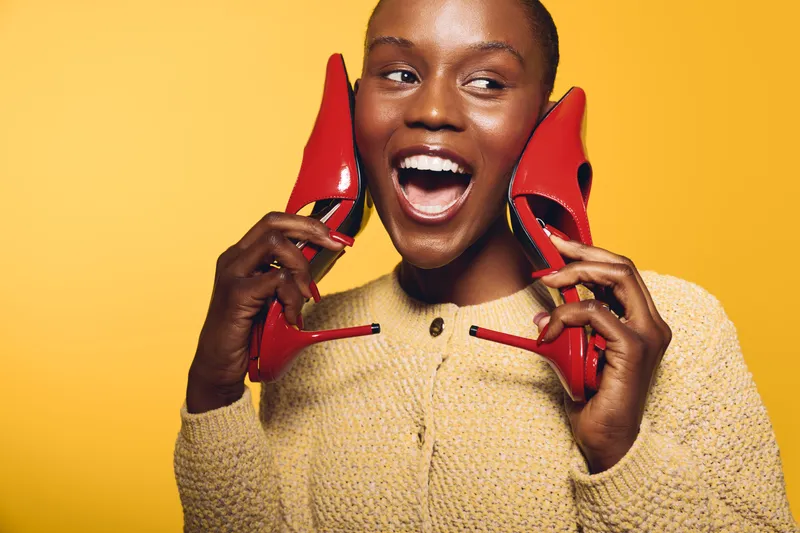 Happy African woman with dark skin holds red high heels near her face smiling broadly on yellow background. Concept of fashion, joy, and beauty with vibrant colors and positive mood.