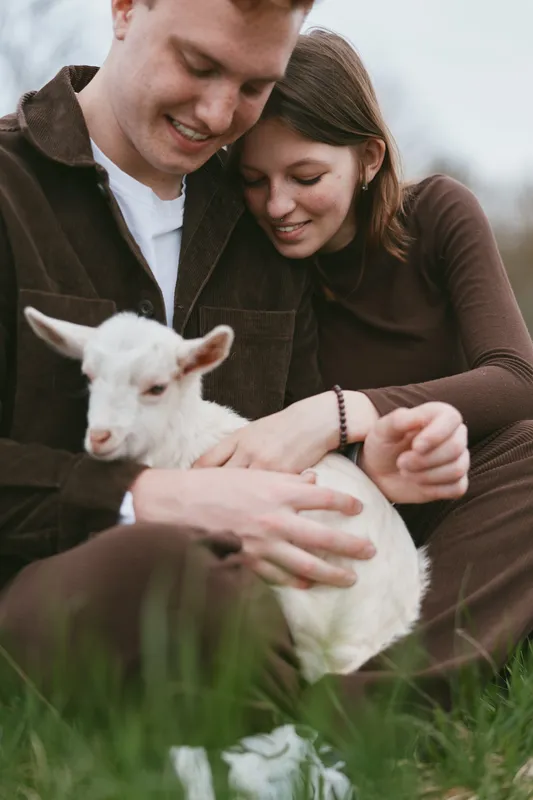 Cheerful couple outdoors with a white goat kid, warm clothes, genuine smiles, sunlit grass meadow, relaxed pose, candid lifestyle moment, natural colors, rustic vibe, playful affection