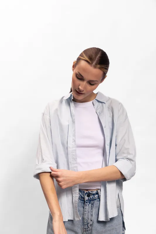 Young woman in casual clothes looks down thoughtfully while adjusting her sleeve against a white background. Concept for introspection, fashion, and relaxed style in studio lighting.