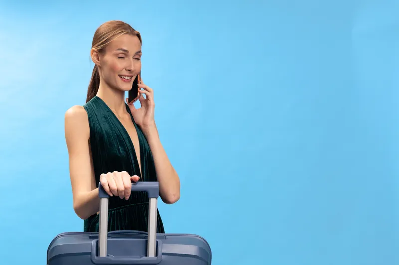 Young woman in green dress holds suitcase and talks on phone, smiling against blue background. Concept of travel, vacation, communication and lifestyle.