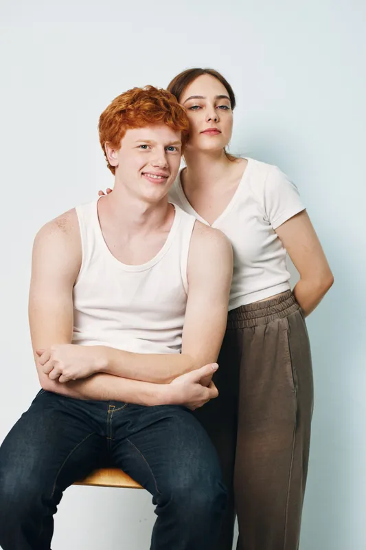 Young man with red hair sitting on stool and smiling with woman standing behind in casual clothes against white background for modern lifestyle portrait.
