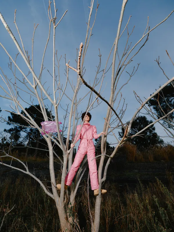 Person wearing pink jumpsuit climbs a bare tree in an open field under daylight, capturing a bold outdoor fashion moment with balance, space, and nature, emphasizing adventure and creative