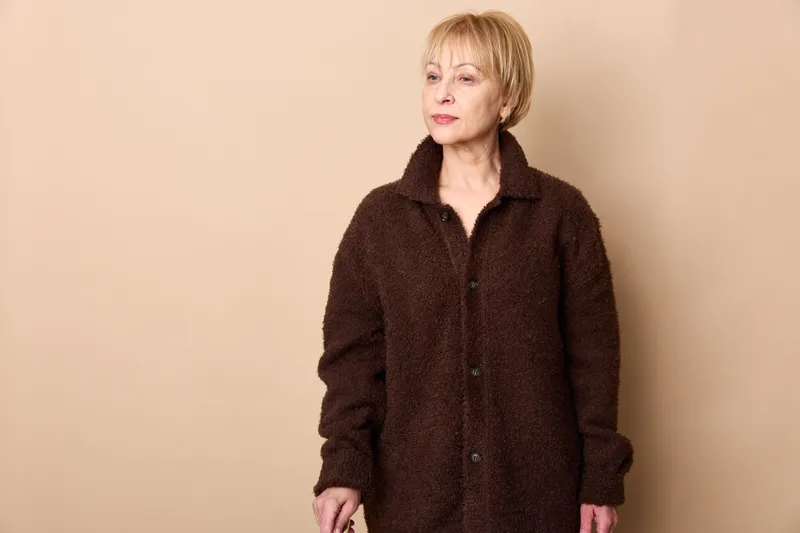Senior blonde woman portrait, wearing dark coat, standing in studio on beige background, calm expression, relaxed pose, minimalist composition, soft natural light.
