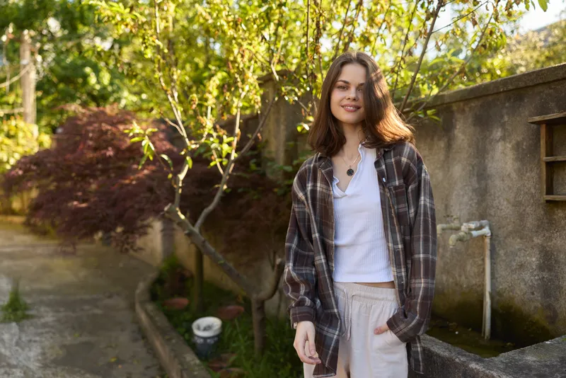 A young woman in a plaid shirt and light trousers stands in a sunlit backyard, smiling softly as a casual lifestyle moment unfolds