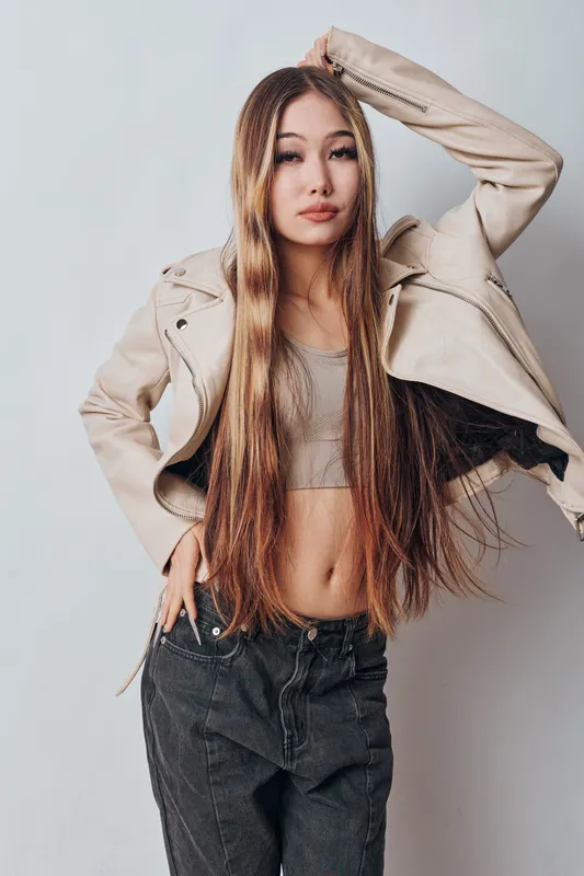 Woman with long hair posing in beige jacket and crop top with black pants against plain background. Fashion, style, confidence and casual look with natural makeup and relaxed posture.