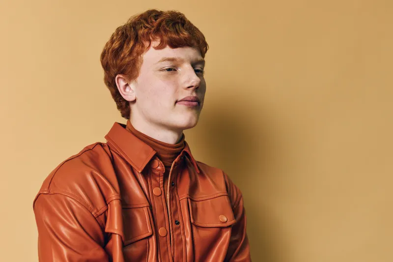 Young man with red hair wearing orange leather jacket and turtleneck sweater poses against beige background for fashion portrait and style concept indoors.