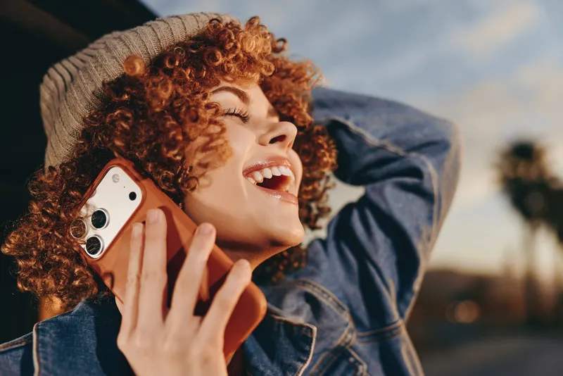 Woman lifestyle outdoors, smiling with curly red hair, denim jacket and knit hat, smartphone in hand, phone call, warm evening light, relaxed mood, city background soft breeze sunlit