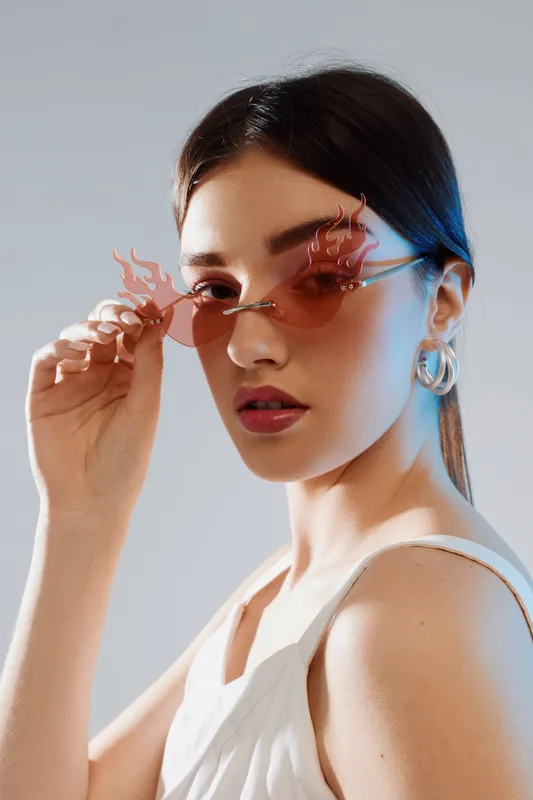 Young woman in white top wearing stylish flame-shaped sunglasses and large hoop earrings poses against neutral background. Concept of woman beauty fashion and modern accessories.