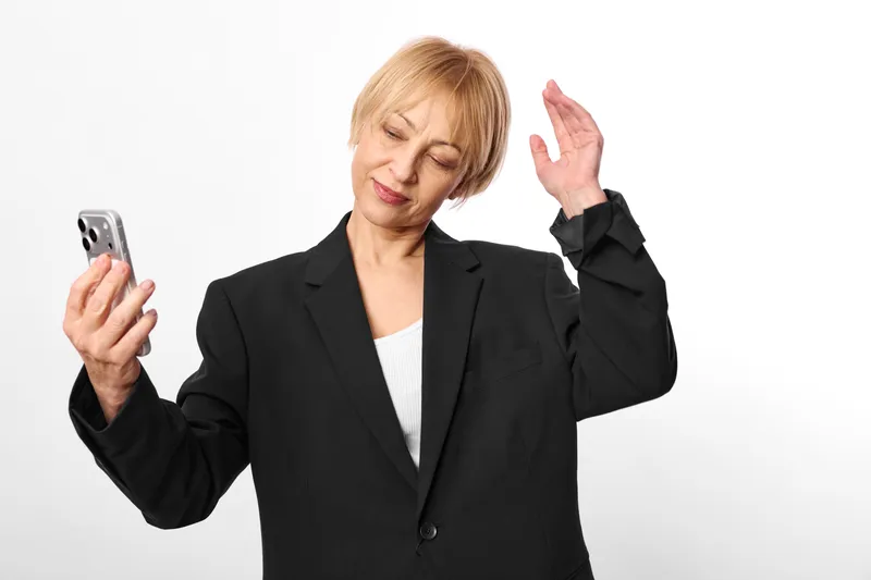Woman in business suit holds smartphone in studio, waving gesture, phone in left hand, annoyed face, isolated on white background, corporate portrait, refusal sign.