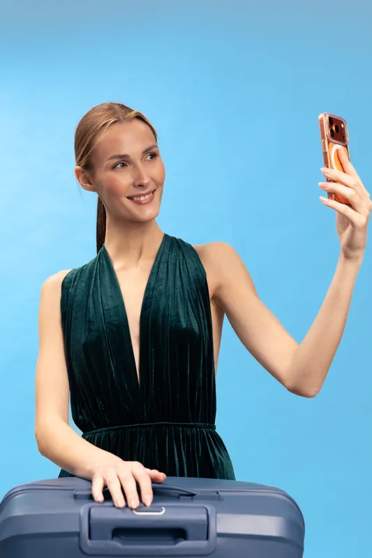 Young woman in elegant dress takes selfie with smartphone while holding suitcase on blue background. Concept of travel, vacation, and modern lifestyle with digital technology and happiness.