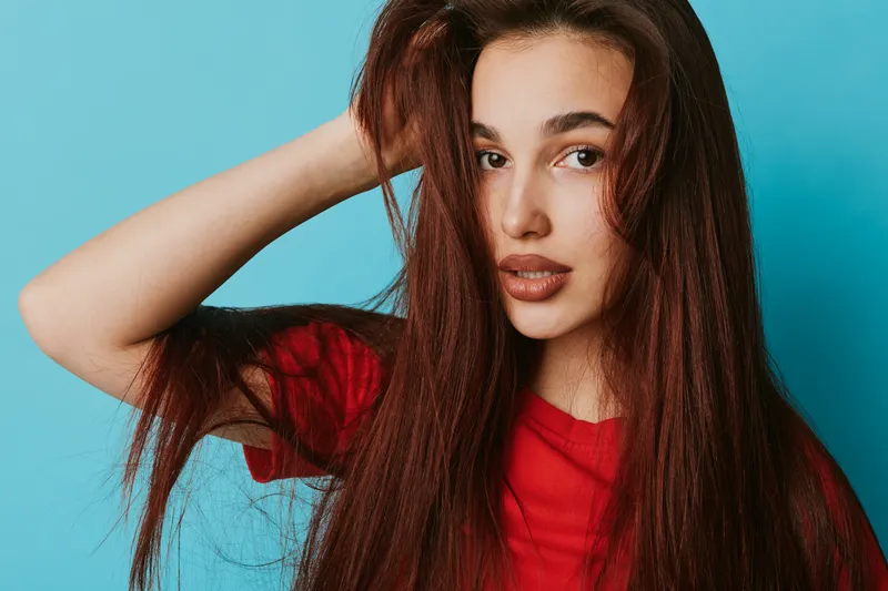 Young woman with long brown hair wearing red shirt posing with hand in hair against blue background for beauty portrait and fashion style. Female model with natural makeup and confident expression.