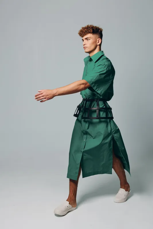 Young man with curly hair wearing green dress and white shoes posing in studio with neutral background. Fashion model in casual outfit with unique belt accessory walking sideways.