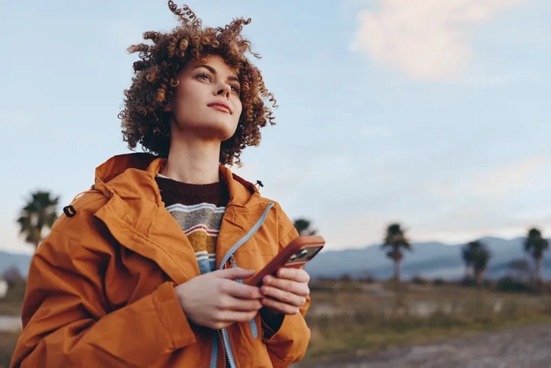 Woman lifestyle portrait outdoors, curly hair and orange jacket, holding smartphone while walking in warm desert landscape, relaxed expression, travel mood, natural light, soft breeze today