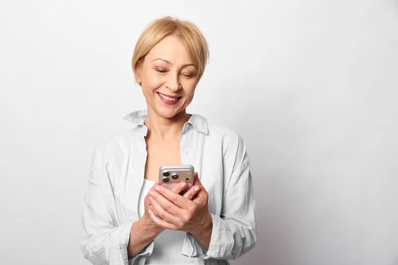 Smiling senior woman holding smartphone in studio portrait, relaxed posture, looking at screen, soft lighting, light gray background, casual shirt, mood, technology communication