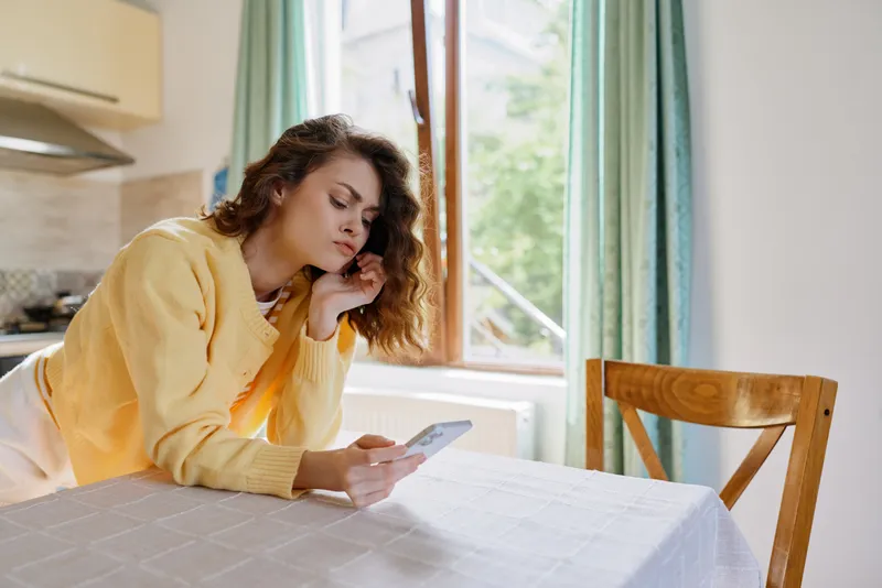 Woman lifestyle home indoors at table using phone, relaxed pose, thoughtful mood, soft natural light, cozy kitchen setting, casual atmosphere, bright window, calm expression, warm colors, everyday