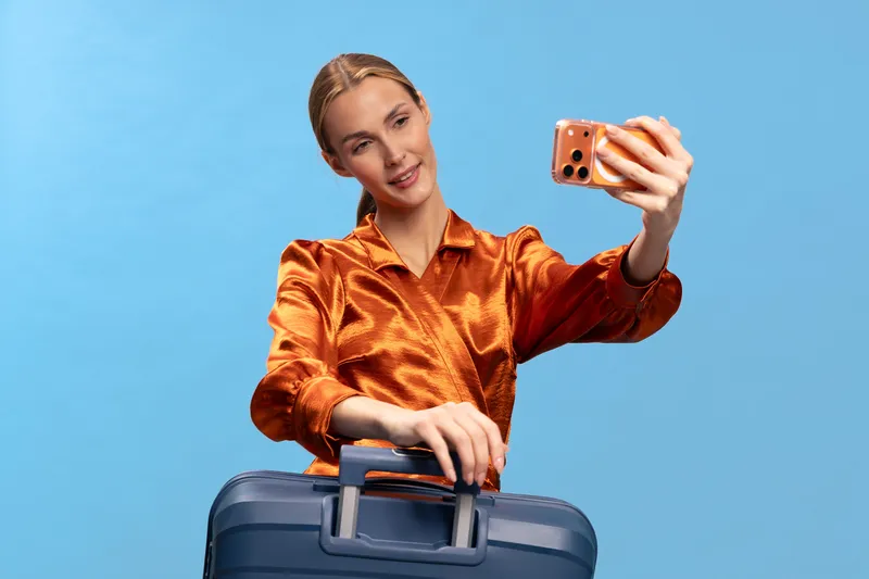 Young woman in orange blouse takes selfie with smartphone while holding blue suitcase handle on blue background. Concept of travel, vacation, tourism, and modern lifestyle.