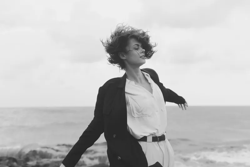 Monochrome image of a woman in a jacket and white shirt on a windy coast by the sea, hair blown, arms outstretched, evoking freedom and motion.