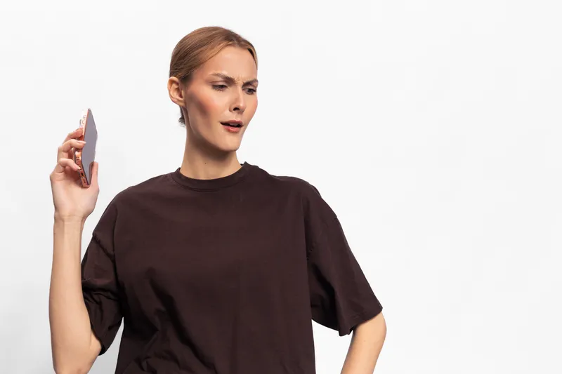 Angry woman with smartphone in hand looks upset and frustrated, standing isolated on white background. Concept of anger, conflict, communication problems, and negative emotions.