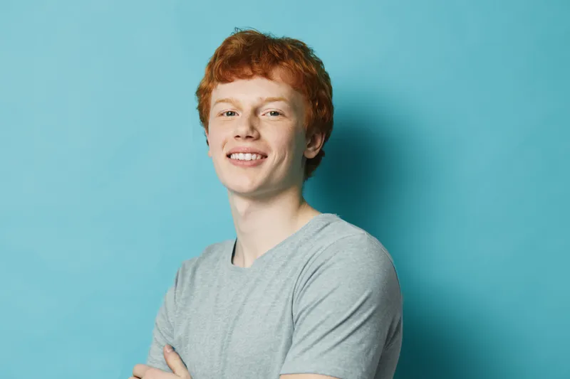 Young man with red hair smiling and crossed arms in casual t shirt against plain blue background. Portrait of confident male with light skin and curly hair studio shot.