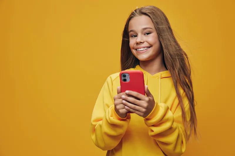 Girl with long hair wearing yellow hoodie smiling and holding red smartphone on yellow background. Young woman using mobile device for communication and technology.