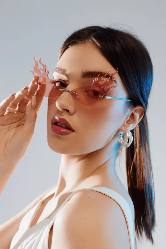 Young woman with long dark hair wearing flame-shaped pink sunglasses and hoop earrings looks confidently at camera. Concept of woman beauty fashion and style.