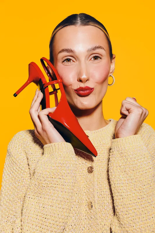 Young woman with red lipstick holds red high heel shoe near face, wearing beige sweater on bright yellow background. Concept of fashion, style, and beauty accessories.